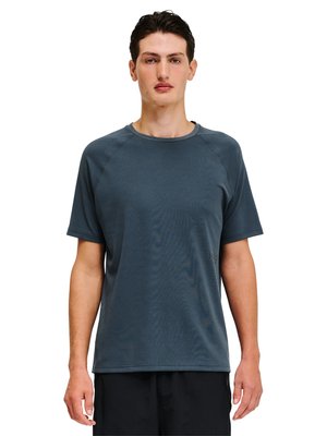 Junger Mann mit kurzen dunklen Haaren, der ein dunkelgraues strukturiertes kurzärmliges T-Shirt und schwarze Hose trägt, steht vor einem weißen Hintergrund.