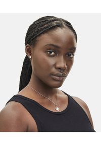 Zilveren halsketting met een vierkante hanger en een ketting, gedragen op een zwarte tanktop. Model heeft gevlochten haar en neutrale make-up, en kijkt recht vooruit.