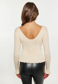 Beige gerippter Strickoberteil mit tiefem V-Ausschnitt hinten, langen Ärmeln und Rüschenbesatz. Kombiniert mit figurbetonten schwarzen Lederhosen.