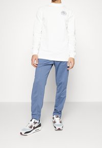 Weißes Langarmhemd mit Logo, blaue Jogginghose und mehrfarbige Sneakers mit einer Mischung aus blauen, weißen und burgunderfarbigen Akzenten.