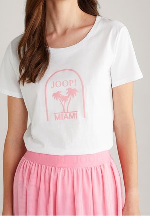 Femme portant un t-shirt blanc avec un motif de palmier rose et le texte "JOOP! MIAMI", assorti à une jupe plissée rose.