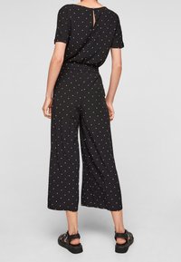 Combinaison noire à pois blancs, manches courtes, taille élastique, coupe jambes larges, et détail œillet dans le dos. Texture de tissu lisse.