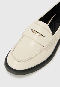 Kreemivärvi nahast loafer ümmarguse varbaga, must rubberist kontsaga ja kuldne logo aktsent rihmal. Omab tikandi detaile.