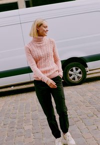 Pink cable-knit turtleneck sweater with ribbed cuffs, paired with black cargo pants and white sneakers, set against a gray van background.