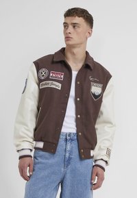 Hnedý varsity kabát s krémovými rukávmi, ktorý obsahuje vyšívané nášivky, kovové cvoky, rebrované manžety a logo na hrudi.