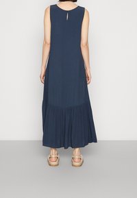 Nainen, joka on pukeutunut ilmanhihaiseen tummansiniseksi maksimekkoon, jossa on kerätty helma, ja beigeihin sandaaleihin, seisoo yksinkertaista vaaleanharmaata taustaa vasten.