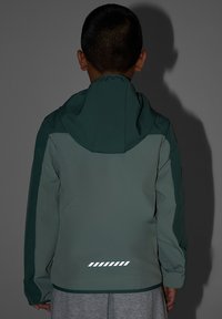 Enfant portant une veste à capuche verte avec des bandes réfléchissantes dans le dos, debout face à un fond gris.