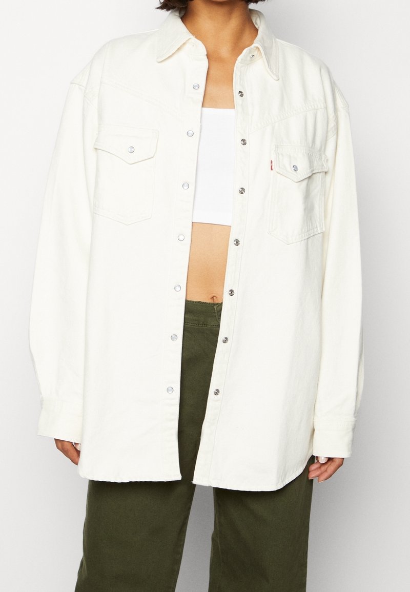 Persona vistiendo una camisa de botón extragrande color blanco roto sobre un crop top blanco y unos pantalones de tiro alto verde oliva, de pie contra un fondo liso.