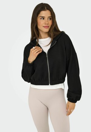 Svart kort jacka med dragkedja och huva, lager på en vit topp. Slätt tyg och avslappnad passform, ihop med beige ribbade leggings.