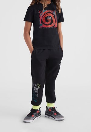 Osoba ubrana w czarny T-shirt z czerwonym wzorem i napisem, czarne spodnie dresowe z kolorowym napisem "O'NEILL", neonowo-żółte skarpetki i szare trampki.