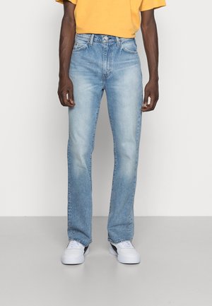 Man wearing light blue straight-leg jeans, white sneakers, and a yellow shirt standing against a plain white background.