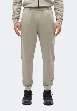 Hombre con pantalones jogger beige de North Face y zapatos de senderismo negros, de pie frente a un fondo claro y liso.