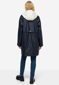 Námořnický voděodolný plášť do deště s bílou kapucí, vybavený zadním větracím otvorem a dekoračními kovovými detaily. Nositelný přes modré džíny a černé boty.