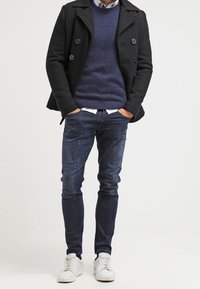 Svart dubbelknäppt sjömansrock över en marinblå tröja, kombinerad med mörk denimjeans och vita sneakers. Outfiten har rena linjer och klassisk design.