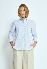 Camicia azzurra chiara a maniche lunghe con un motivo a diamante testurizzato, colletto con bottoni e orlo sfilacciato. Abbinata a pantaloni ampi color beige chiaro.