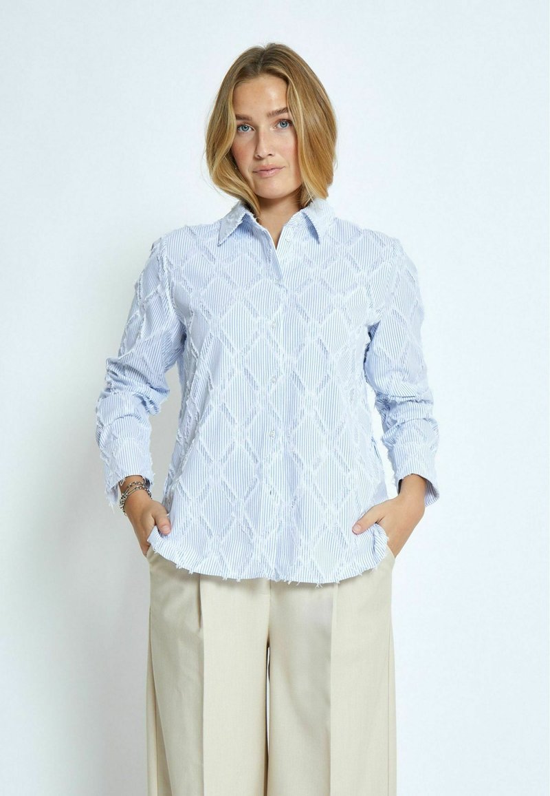Camicia azzurra chiara a maniche lunghe con un motivo a diamante testurizzato, colletto con bottoni e orlo sfilacciato. Abbinata a pantaloni ampi color beige chiaro.