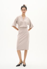 Knielanger, figurbetonter Rock aus weichem beigen Stoff; kurzes, weites Top mit V-Ausschnitt. Ein Paar schwarze Slingback-Absätze rundet den Look ab.