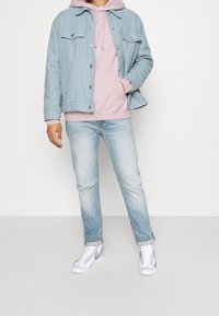 Chaqueta de mezclilla azul claro con botones y dos bolsillos en el pecho, sobre una sudadera rosa. Jeans azul claro y zapatillas blancas con acentos grises.