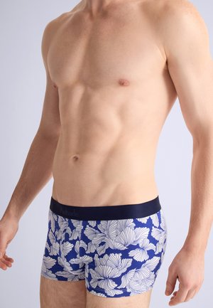 Homme torse nu portant un boxer bleu avec un motif floral blanc, debout les bras détendus sur un fond clair uni.