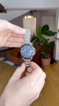 RVS-horloge met een blauwe wijzerplaat, zilverkleurige uurmarkeringen, zwarte bezel en chronograaf functies. Armband met schakels.