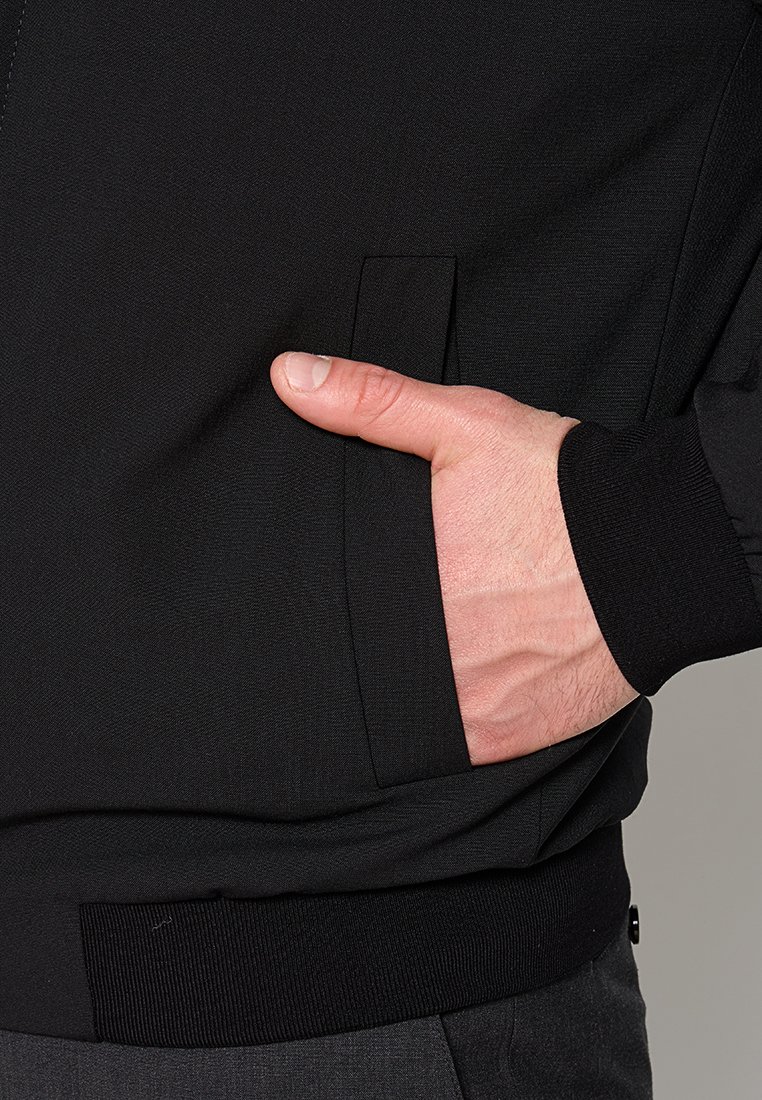 Schwarze Jacke mit glatter Textur, ausgestattet mit einer seitlichen Tasche und gerippten Bündchen. Minimalistisches Design mit einer taillierten Silhouette.