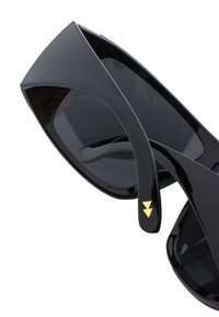 Mustat aurinkolasit kiiltävällä pinnalla, ylisuurilla neliömäisillä kehyksillä, tummilla laseilla ja kultaisilla kolmiomaisilla yksityiskohdilla temppeleissä.