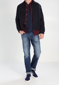 Marinblå mockajacka över en vinröd knappad skjorta, ihop med blå jeans och marinblå sneakers. Detaljer inkluderar två framfickor och uppvickade ärmar.