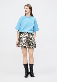 Giovane donna che indossa una maglietta blu larga, una minigonna leopardata con tasche e stivali neri fino a metà polpaccio, in piedi davanti a uno sfondo semplice.