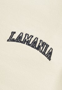 Siuvinėjimas ant šviesiai smėlio spalvos audinio pavaizduoja žodį "LAMANIA" tamsiai mėlyna spalva, su nežymiai pakelta tekstūra ir išlinkusiu dizainu.