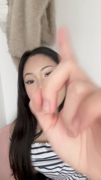 Jeune femme aux longs cheveux bruns pointant du doigt près de la caméra, manteau en fourrure beige accroché à un mur blanc derrière elle.