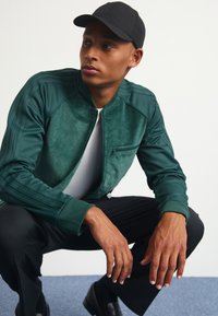 Mörkgrön bomberjacka med en texturerad framdel, ribbade ärmslut och en ficka med dragkedja, tillsammans med svarta byxor och en svart keps.