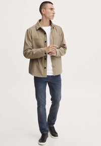 Beige Flanell-Hemdjacke mit zwei Brusttaschen, knöpfbarem Vorderteil, über einem weißen T-Shirt getragen, kombiniert mit blauen Jeans und schwarzen Sneakers.