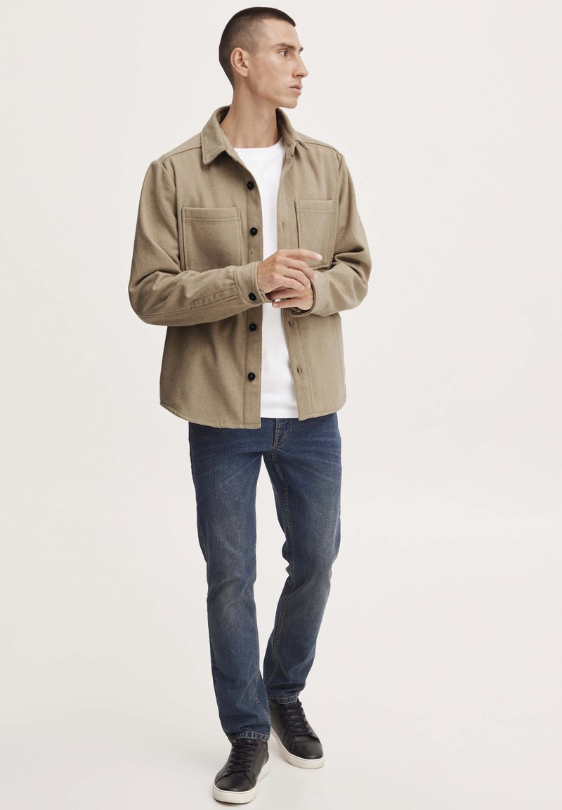 Beige Flanell-Hemdjacke mit zwei Brusttaschen, knöpfbarem Vorderteil, über einem weißen T-Shirt getragen, kombiniert mit blauen Jeans und schwarzen Sneakers.