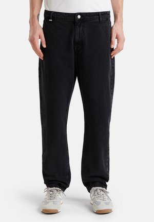 Pantalones de mezclilla negros con un corte relajado, que presentan un cierre de botón, bolsillos laterales y un leve desvanecimiento. Combinados con zapatillas de color claro.