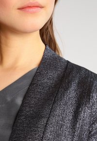 Americana negra texturizada con un acabado metálico, con un amplio solapa y una blusa gris suave debajo.