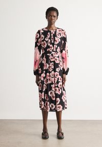 Mujer vistiendo un vestido midi negro con mangas largas y estampado floral en rosa, de pie sobre un suelo gris contra una pared blanca.