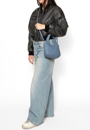 Femme portant une veste en cuir noire, un jean bleu clair à jambes larges, des baskets blanches, tenant un petit sac à main bleu avec une bandoulière.