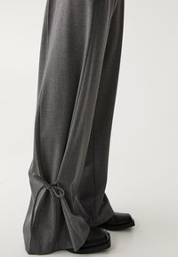 Pantalons gris ajustés en tissu texturé, dotés d'une coupe évasée et d'accents noués aux ourlets, accompagnés de chaussures noires.