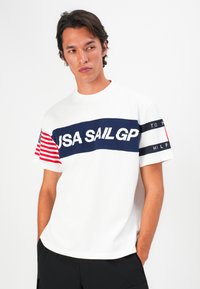 Tânăr cu păr închis la culoare, îmbrăcat cu un tricou alb pe care scrie "USA SAIL GP", cu steagul american pe un mânecă și cu pantaloni scurți negri, stând în fața unui fundal alb.