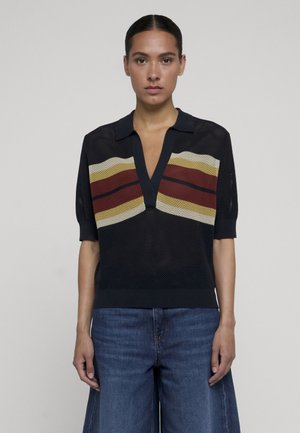 Frau mit dunklem, zurückgebundenem Haar, die einen schwarzen, kurzärmeligen Stricktop mit roten, gelben und beigen Streifen trägt, kombiniert mit blauen Jeans mit weitem Bein.