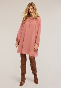 Donna che indossa un vestito lungo a maniche lunghe di un rosa polveroso, con maniche plissettate e stivali di pelle marroni al ginocchio, in piedi di fronte a uno sfondo semplice.