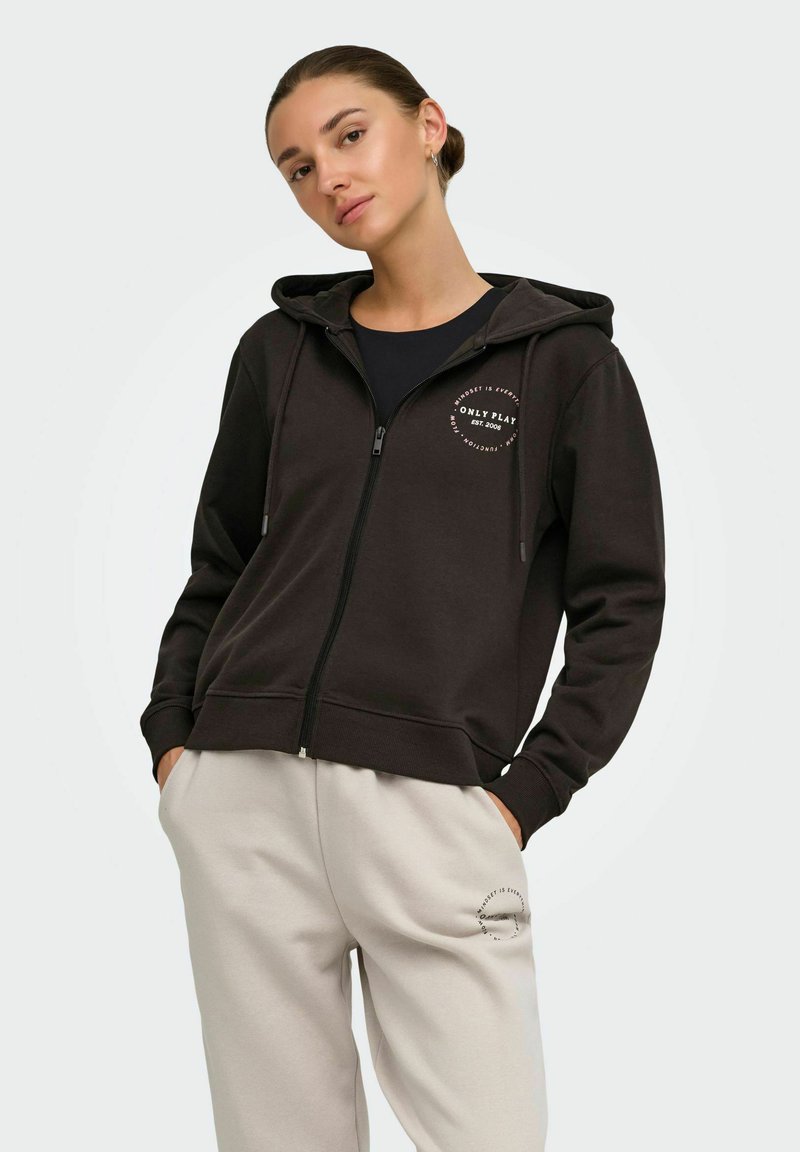 Femme portant un sweat à capuche noir zippé avec capuche et pantalon de jogging beige, debout les mains dans les poches devant un fond uni.