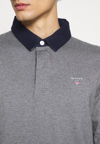 Graues Baumwoll-Polohemd mit navyfarbigem Kragen, versehen mit einer Knopfleiste und besticktem GANT-Logo auf der Brust. Glatte Textur.