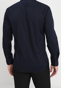 Hombre vestido con una camisa azul marino de botón de manga larga ajustada y pantalones negros, visto desde atrás contra un fondo liso.