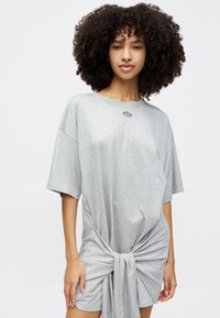Siva, oversize T-shirt obleka iz mehkega bombaža z kratkimi rokavi in zavezanim detajlom na pasu. Ima majhen srebrn poudarek pri ovratniku.