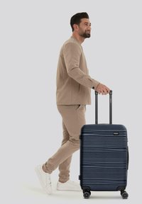 Maleta rígida navy con diseño de surcos horizontales, mango retráctil y cuatro ruedas, siendo arrastrada junto a una persona vestida de beige.