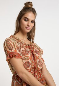 Blusa a motivi floreali in beige, con maniche volanti, scollo rotondo e dettagli con cordoncino. Accenti prominenti in rosso e arancione.