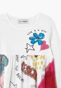 Biała sukienka z długim rękawem z niebieskimi kwiatami i różowymi sercami, z kolorową, wzorzystą spódnicą w różnych odcieniach i fakturach.