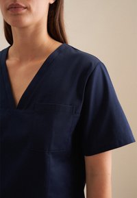 Maglietta da scrub blu navy con scollatura a V, maniche corte e taschino sul petto. Il tessuto sembra liscio e leggero.
