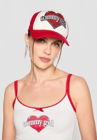 Red and white trucker cap featuring a glittery heart design and "tommy girl" text, with a mesh back and curved brim.